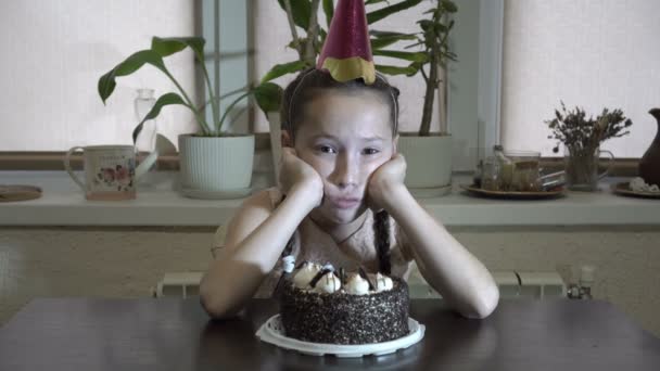 Menina triste sentada à mesa com a cabeça apoiada com as mãos com um boné festivo na cabeça e olha para a câmera. Antes dela em cima da mesa é um bolo. Fecha. 4K. 25 fps — Vídeo de Stock
