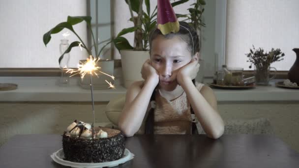 A sad child, a little girl, sits at the table and looks at the burning Sparkler and the cake that is on the table. She sits supporting his head in his hands. Close up. 4K. 25 fps — Stock Video