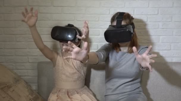 As crianças jogam jogos virtuais. Duas irmãs em óculos de realidade virtual sentam-se e brincam em casa no sofá contra uma parede de tijolo branco. Acenam as mãos e olham em volta. Fecha. 4K. 25 fps — Vídeo de Stock