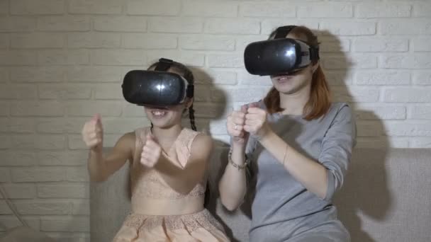 En liten flicka och hennes tonåriga syster i virtual reality-glasögon sitter och spelar virtuella spel hemma i soffan på en vit bakgrund. De viftar med händerna och titta runt. — Stockvideo