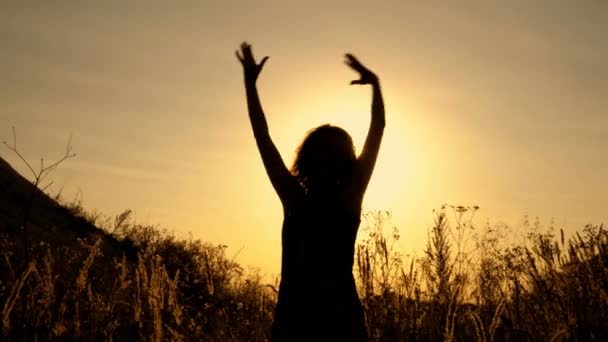 女孩在明亮的阳光日落的背景下站着 在山间田野里的花丛中挥手致意 底部视图 闭上你的嘴4K — 图库视频影像