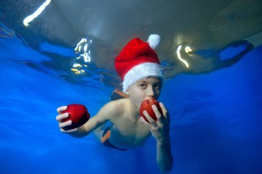 Portrait of a little boy who bites a red Apple underwater in the pool in a red Santa hat on a blue background. Conceptual realism. Portrait. Landscape orientation of the image. clipart