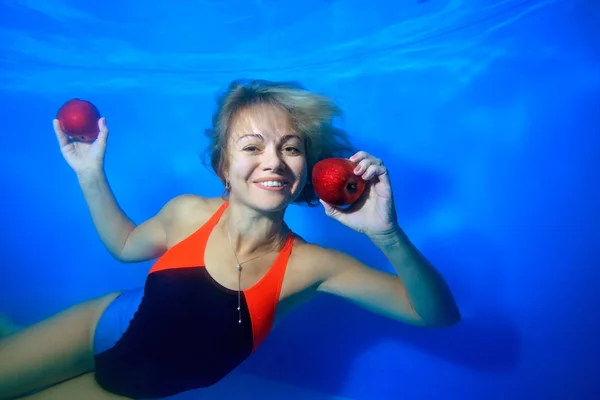 Happy young girl smiling and floating underwater in the pool near the bottom with red apples in her hands on a blue background and looking at the camera. Portrait. Concept. Landscape orientation.