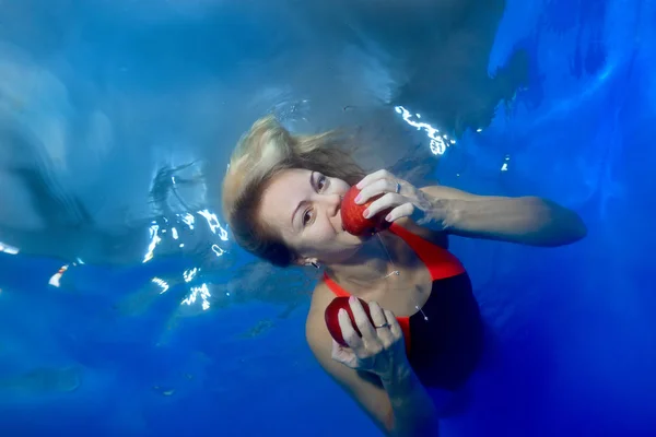 Underwater portrait of a cute girl with red apples in her hands on a blue background. She swims underwater with disheveled hair, looks at the camera and bites an apple. Conceptual realism.