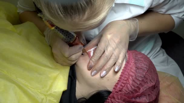 在美容院里 一个女人做了一个下唇的纹身 一个专业的美容师用一根针把油漆涂在女人的嘴唇上 闭上你的嘴从顶部的视图 — 图库视频影像