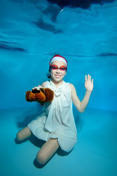 A child with a toy-dog in his hand sitting at the bottom of the pool under the water in a white dress and swimming goggles, smiling and waving on a blue background. Vertical orientation of the image.