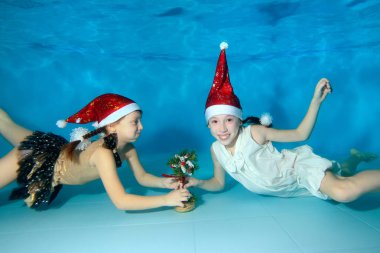 Noel Baba'nın kırmızı şapka çocuklarda yüzmek ve mavi bir arka plan üzerinde küçük bir Noel ağacı ile su altı oyun, kamera ve gülümseme bak. Portre. Sualtı çekim. Manzaralı.