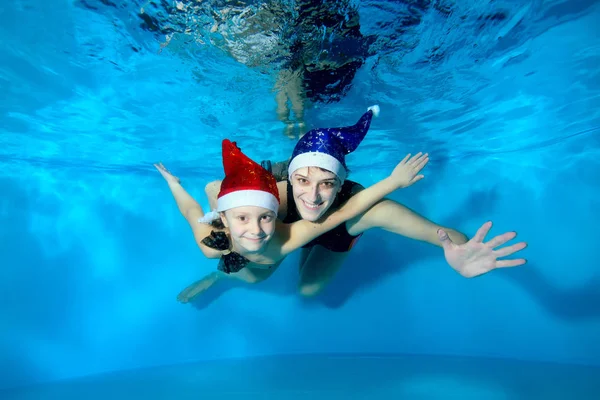Mom with a little daughter swim and play underwater in the pool on a blue background in Santa\'s hats. They look at the camera and smile. Portrait. Landscape orientation.