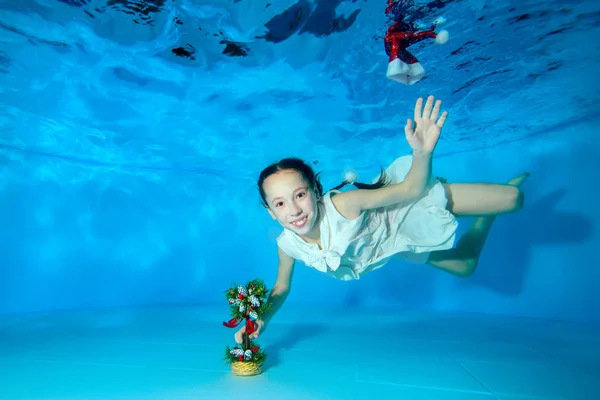 Happy little girl underwater swims to the Christmas tree, looks at the camera and smiles. Portrait. Shooting underwater. Landscape view.