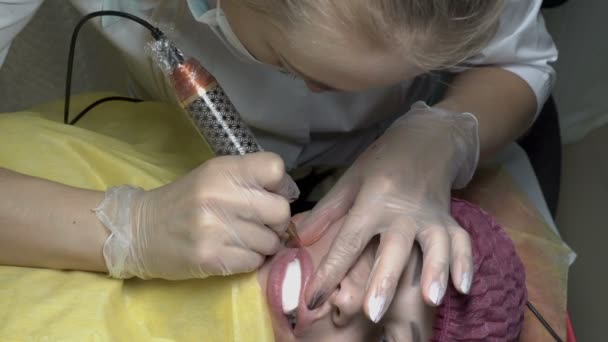 在美容院永久化妆嘴唇的过程中 女性美容师用针头和油漆治疗女孩的嘴唇 从顶部的视图 闭上你的嘴4K — 图库视频影像