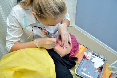 Beyaz bir tıbbi elbise güzellik uzmanı Avrupalı bir Bayan kalıcı yapar dudak makyaj güzellik salonu ve ona bir iğne dudakları ile pembe boya koyar. Üstten görünüm. Yatay yönlendirme.