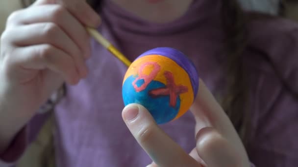 Une petite fille tient un œuf de Pâques dans sa main et peint dessus un pinceau et des peintures colorées, assise à la maison à la table. Ferme là. 4K. 25 ips — Video