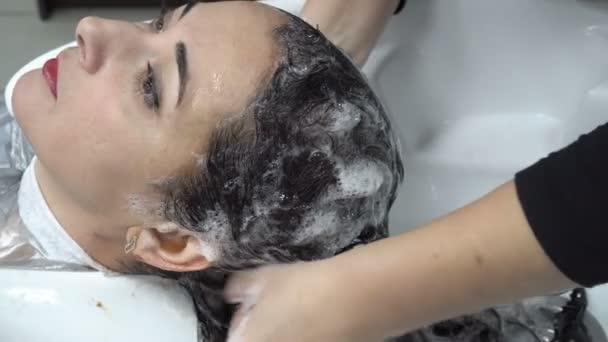 Крупным планом стилист вымывает волосы восточной женщины в парикмахерской над белой раковиной. Уход за волосами. 4К. 25 кадров в секунду — стоковое видео