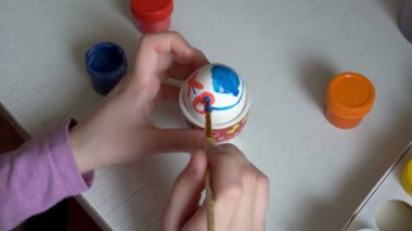 Çocuk tatil hazırlanma ve masada oturan mavi ve kırmızı boya Paskalya yortusu yumurta sembolleri çizer. Yakın çekim. Üstten Görünüm. 4 k. 25 fps