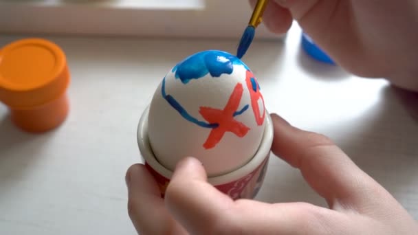 Чрезвычайно крупным планом ребенок рисует пасхальное яйцо сине-красной краской, сидя за столом. 4К. 25 кадров в секунду — стоковое видео
