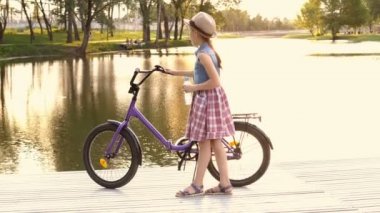 Gün batımında nehir kıyısında küçük bir kız bisikleti için gelir ve şişeden su içmek başlar. Parlak sarı günbatımı. Portre. 4 k. 25 fps.