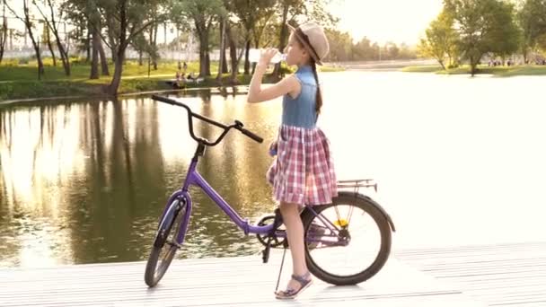 Una niña sentada en una bicicleta lila cerca del río al atardecer y bebiendo agua de una botella transparente. Atardecer amarillo brillante. Retrato. 4K. 25 fps . — Vídeo de stock