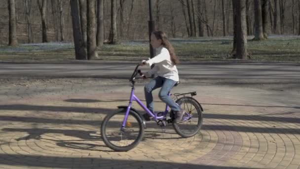 Una niña montado en un círculo en una bicicleta lila en un parque de primavera en ropa blanca al atardecer. Parque municipal. 4K. 25 fps . — Vídeo de stock