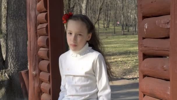 Schattig klein meisje met haar haren naar beneden poseren op de speelplaats in het voorjaar Park. Ze zit op een houten bankje en kijkt naar de camera. Portret. Close-up. 4 k. 25 fps. — Stockvideo