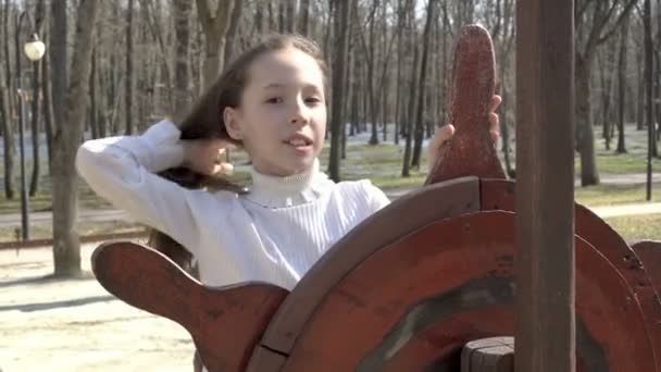 La bambina si raddrizza svolazzando tra i capelli del vento, in piedi al timone della nave sul campo da giuoco nel parco primaverile. Guarda la telecamera e sorride. 4K. 25 fps . — Video Stock