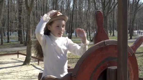 Una joven se para y juega en un bote de madera al timón de un parque infantil en el parque. Se quita el sombrero, le saluda, mira a la cámara y sonríe. Cabello revoloteando en el viento. 4K . — Vídeos de Stock