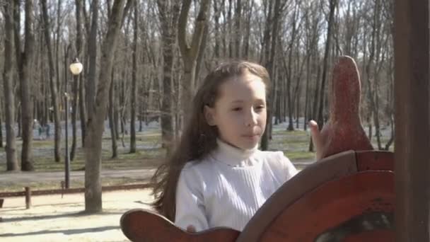 Een klein meisje speelt kapitein aan het roer van een houten schip op de speelplaats in het Spring Park. Ze rechtzetten haar haar, fladderen in de wind, kijkt naar de camera en glimlacht. 4k. 25 fps. — Stockvideo