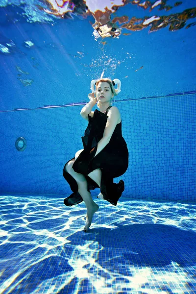 Mooie blonde meisje poseren onder water in het zwembad in een zwarte jurk en met witte hoorns op haar hoofd op een zonnige dag. Portret. Cosplay. Fotograferen onderwater. Verticale afdrukstand van de afbeelding — Stockfoto