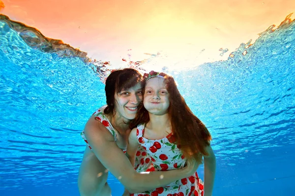 Sonrientes madre e hija nadan y posan bajo el agua en la piscina contra la puesta de sol naranja brillante en los mismos vestidos. Retrato. Disparando bajo el agua. Orientación horizontal de la imagen — Foto de Stock