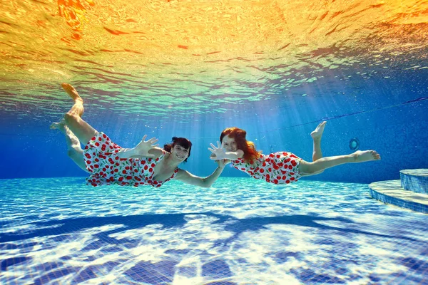 Mamãe e filha nadam um para o outro sob a água na piscina contra o pôr do sol tropical brilhante. Eles olham para a câmera e sorriem. Retrato. Tiro debaixo d 'água a partir do fundo — Fotografia de Stock