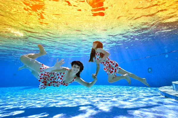 Сім'я, мати і маленька дочка плавають і грають під водою в басейні в одязі на тлі яскравого тропічного заходу сонця. Мама дивиться на камеру і посміхається. Портрет — стокове фото