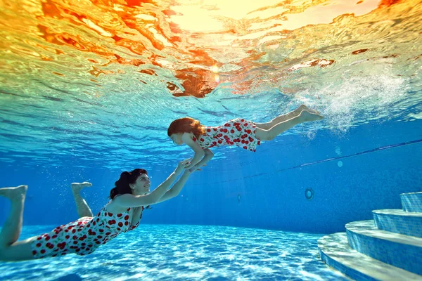 Una niña se sumerge con su madre en el fondo de la piscina sobre el fondo de una brillante puesta de sol tropical. Mamá sonríe y toma las manos de su hija. Retrato. Disparos bajo el agua desde el fondo —  Fotos de Stock