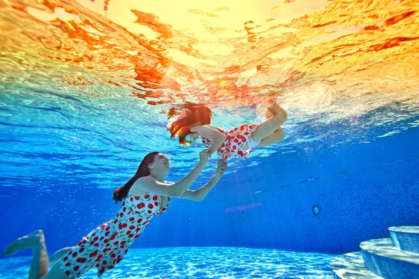Mom teaches her little daughter to swim and dive, and plays with her under the water in the pool against the bright tropical sunset. Mom smiles and supports the daughter\'s hands. Portrait