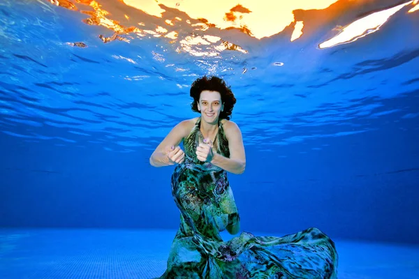 Cute girl in a fashionable dress posing underwater in the pool on a blue background. She looks at the camera and smiles. Portrait. Shooting underwater — Stock Photo, Image
