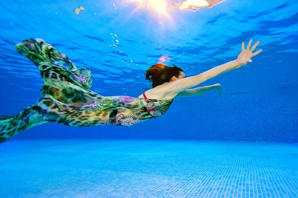 Modelo menina nada e posando debaixo d 'água na piscina em um vestido em um fundo azul. A disparar debaixo de água. Vista lateral. Orientação horizontal da foto — Fotografia de Stock