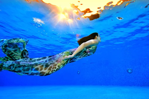 Une fille en robe nage sous l'eau du fond de la piscine à la surface de la lumière du soleil. Tirer sous l'eau. Vue latérale. Orientation horizontale de la photo — Photo