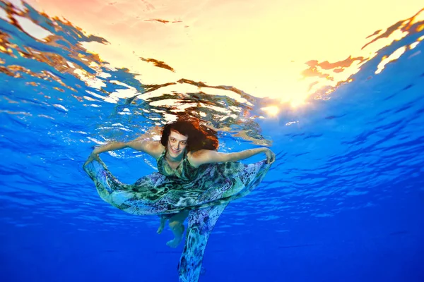 Una chica feliz en un hermoso vestido nada bajo el agua en la piscina contra el fondo de la luz del sol en la superficie, mira a la cámara y sonríe. Retrato. Disparando bajo el agua. Vista horizontal — Foto de Stock