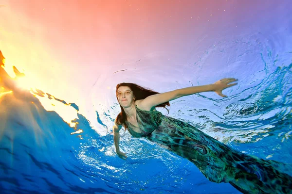 Een mooi model in een jurk zwemt onder het water in het zwembad tegen de achtergrond van zonlicht op het oppervlak, gekeken naar de camera en glimlach verspreiding van zijn handen op de zijkanten. Portret — Stockfoto