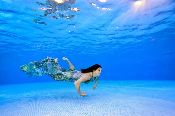 Chica nada en un vestido bajo el agua cerca del fondo de la piscina sobre un fondo azul y sonríe. Retrato. Disparando bajo el agua. Vista del paisaje — Foto de Stock