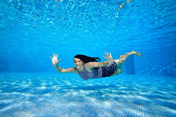Una chica deportiva alegre nada bajo el agua cerca del fondo de la piscina en un traje de baño a rayas en un día soleado brillante, mira a la cámara, agita las manos y sonríe. Retrato. Fotografía submarina — Foto de Stock