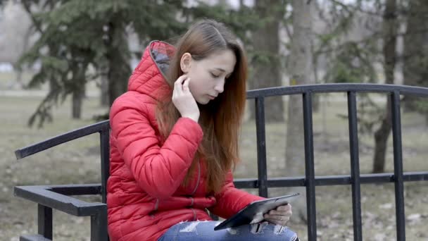 Una joven con una tableta digital en las manos sentada en un parque de primavera en un banco con una chaqueta roja, endereza su cabello y mira la tableta. Retrato. De cerca. 29,97 fps . — Vídeos de Stock