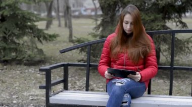 Kırmızı saçlı, kırmızı bir ceket içinde bahar parkta bir bankta oturan bir dijital tablet tutarak ve onu leafing ile düşünceli kız. Portre. RAW video. 29,97 kare/sn fps.