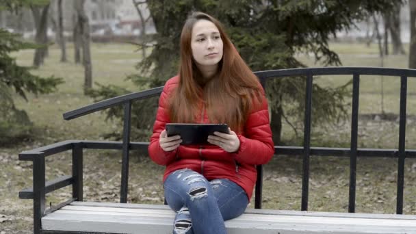 Söt flicka med rött hår innehar en digital tablett och bläddrande genom det, sittande på en bänk i parken på våren i en röd jacka. Porträtt. Närbild. RAW-video. 29,97 fps. — Stockvideo