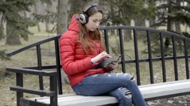 Kırmızı bir ceket bir bahar Park bir bankta kulaklıklar oturan ve bir tablet üzerinde çalışan bir genç kız. Kızıl saçlı kız. Portre. RAW video. 29,97 kare/sn fps.