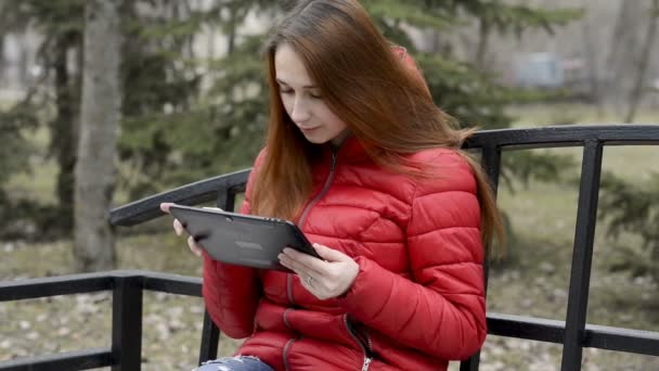 In Großaufnahme sitzt ein junges Mädchen mit roten Haaren in roter Jacke auf einer Parkbank, hält ein digitales Tablet in der Hand und blättert darin. Porträt. Rohvideo. 29,97 fps. — Stockvideo