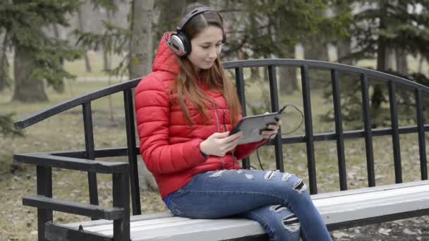 Chica con auriculares en un banco en el parque de primavera sonríe y se ríe mirando la pantalla de la tableta, que se sostiene. Una chica con el pelo rojo en una chaqueta roja. Retrato. Vídeo crudo. 29,97 fps . — Vídeo de stock
