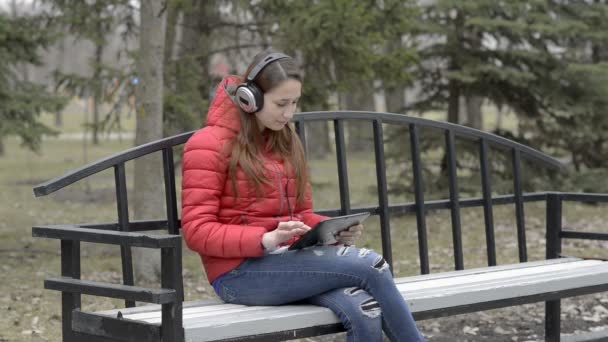 Una joven con auriculares grandes escuchando música sentada en un banco en el parque de primavera. Ella sonríe y asiente a la música, mirando la pantalla de la tableta que sostiene. Retrato. 29,97 fps . — Vídeos de Stock