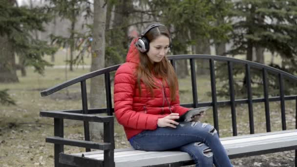 可爱的女孩坐在长凳上, 头上戴着大耳机, 在空荡荡的城市公园里听音乐和唱歌。她微笑着环顾四周。从左到右平移。肖像。29.97 fps. — 图库视频影像