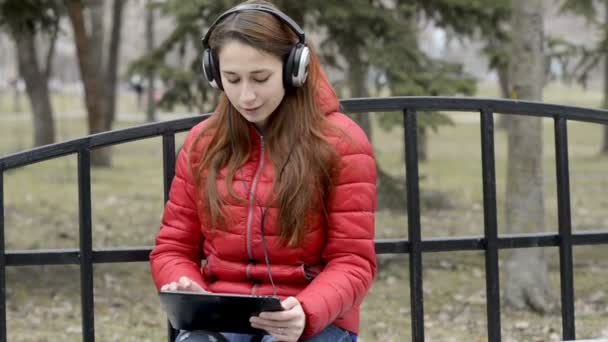 Une adolescente écoute de la musique dans de grands écouteurs et danse sur son rythme, assise sur un banc dans le parc de la ville. Elle regarde l'écran de la tablette qu'elle tient. Panorama de droite à gauche . — Video