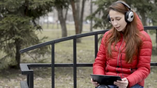 Una buena chica con una tableta y grandes auriculares en la cabeza escucha música y baila a su ritmo, sentado en un banco en el parque de primavera de la ciudad. Panorama de izquierda a derecha. De cerca. 29,97 fps . — Vídeo de stock