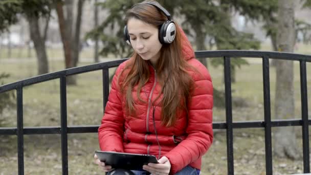 Una chica con una tableta en las manos y auriculares grandes en la cabeza está sentada en un banco en el parque de primavera de la ciudad y escuchando música. Lleva una chaqueta roja y pelo rojo. De cerca. 29,97 fps . — Vídeo de stock